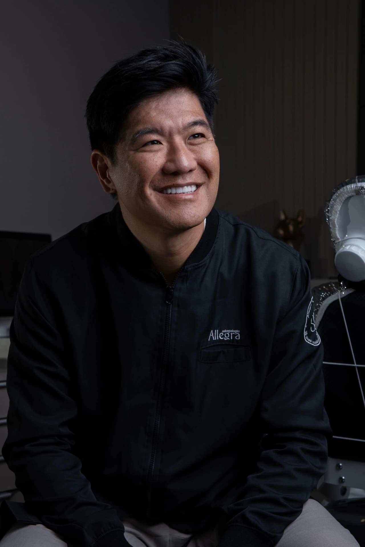 Dr. Rodolfo Segawa sentado em ambiente clínico, sorrindo e usando uniforme preto da Allegra.