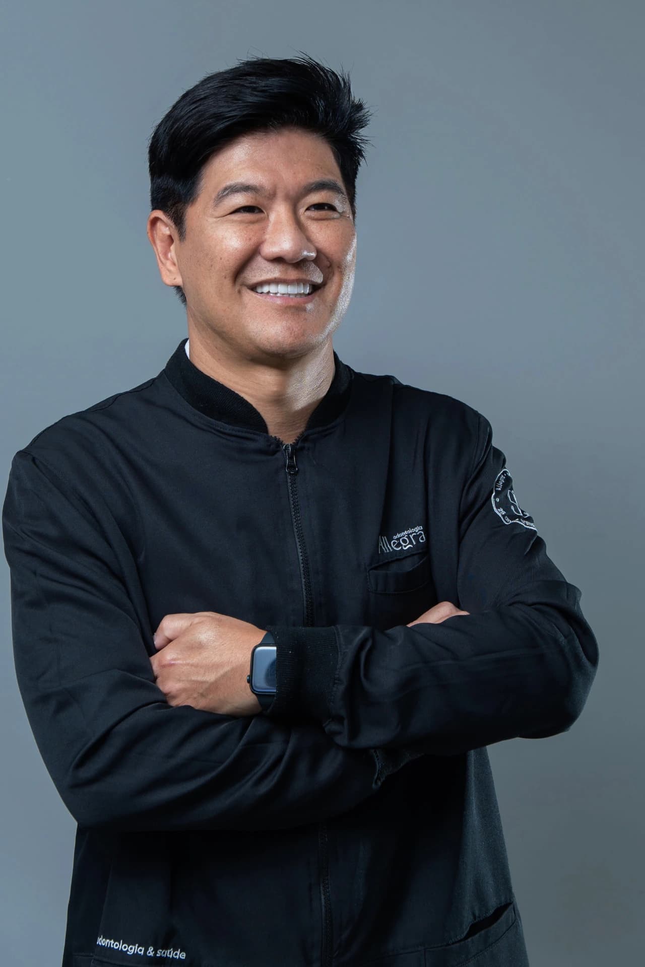 Dr. Rodolfo Segawa de braços cruzados, em uniforme clínico preto, diante de fundo cinza.